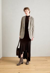 Blazer de cuadros en beige y negro, combinado con un vestido ajustado marrón oscuro con dobladillo con volantes, botas de tobillo grises y un bolso cruzado negro.