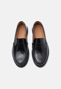 Melnu spīdīgu ādas loafers zābaku pāris ar noapaļotiem pirkstiem un izšuvumu, skatīts no augšas uz gaiša fona.