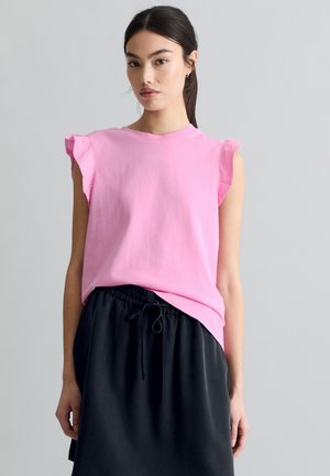 Jeune femme aux cheveux foncés attachés en queue de cheval, portant un haut rose à manches à volants et une jupe noire à cordon, debout devant un fond gris uni.