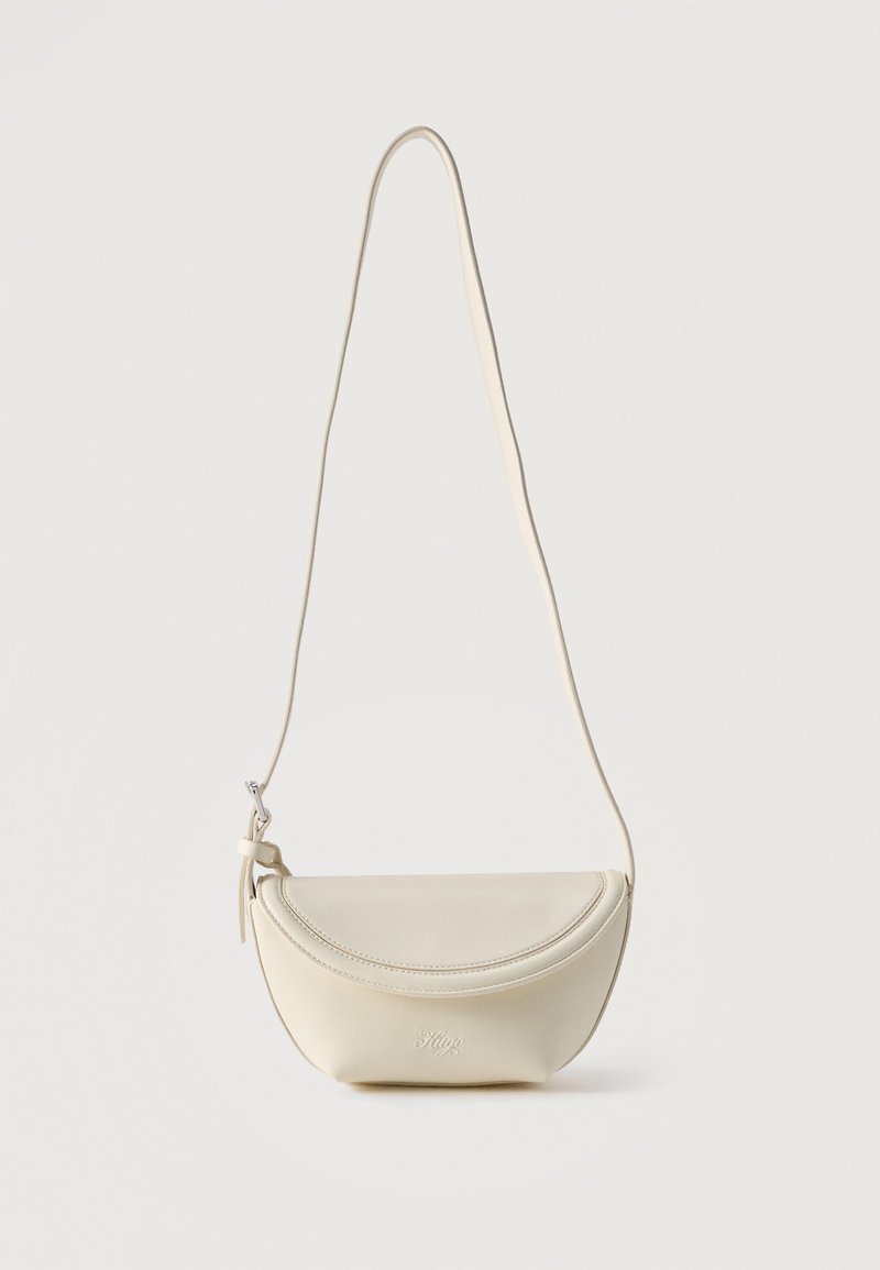 Μικρό δερμάτινο τσαντάκι χρώματος κρεμ, με καμπύλο καπάκι και μακρύ λεπτό λουράκι, κρεμασμένο σε απλό φόντο.