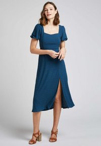 Robe midi texturée de couleur bleu canard avec un décolleté carré, des manches bouffantes et une fente sur le côté, associée à des sandales à talons marron.