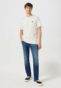 Camiseta de color beige claro con un pequeño logotipo negro, combinada con jeans rectos azules y zapatillas negras con suelas blancas. Corte holgado y diseño simple.