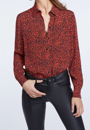 Femme portant une chemise boutonnée à imprimé léopard rouge et noir rentrée dans un pantalon en cuir noir taille haute, avec une main dans une poche.