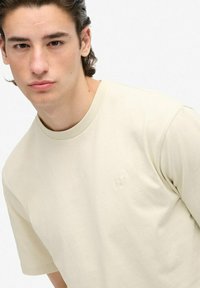 Camiseta de algodón color crema con cuello redondo y mangas cortas. Presenta un discreto logo bordado en el pecho.