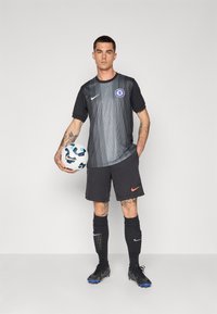 Zwart en grijs voetbalshirt met verticale strepen, voorzien van het Chelsea-logo, gecombineerd met zwarte shorts en zwarte voetbalschoenen. Houdt een voetbal vast.