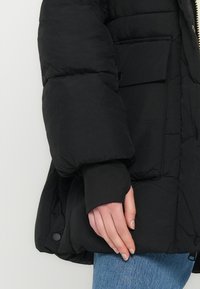 Hånd hvilende ved siden iført en svart dunjakke med elastisk mansjett, kombinert med blå denimjeans mot en nøytral bakgrunn.