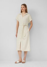 Abito in lino beige chiaro con colletto, maniche corte arrotolate, bottoni frontali, vita con cinturino e spacco fino al ginocchio. Indossato con sandali piatti.