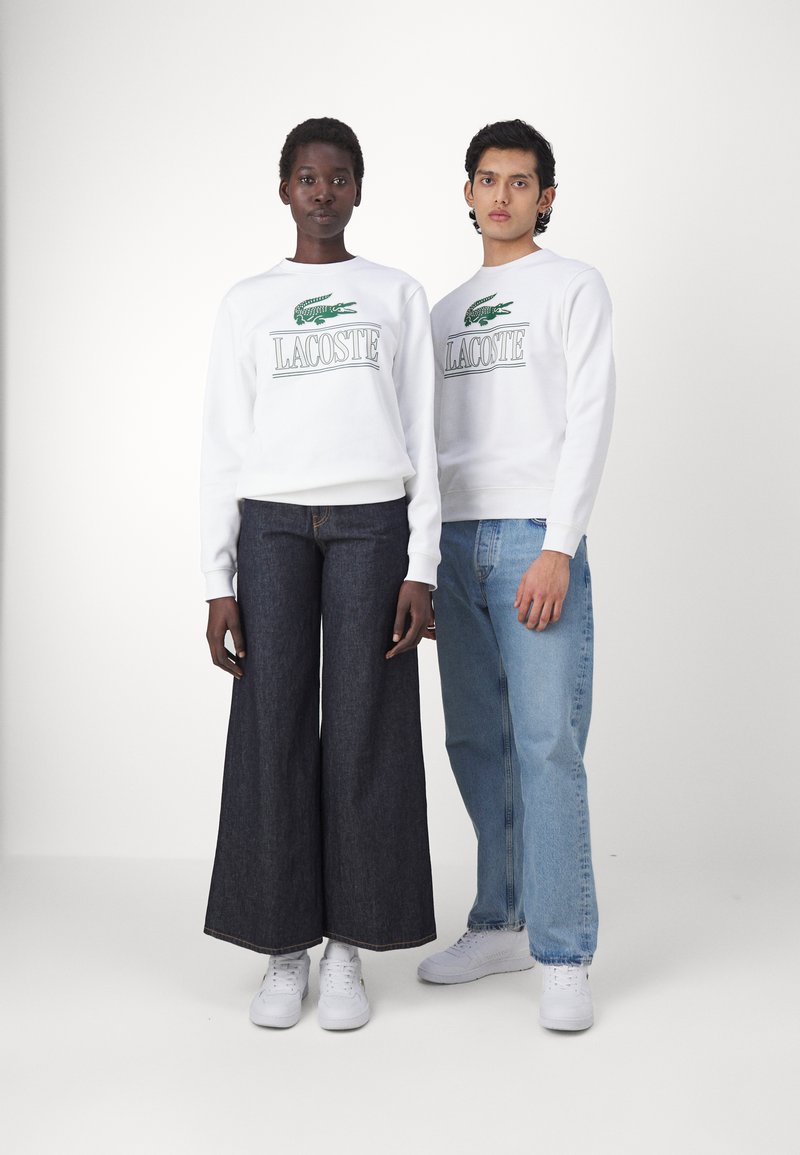 Sudaderas blancas con el logo verde "LACOSTE" combinadas con jeans anchos oscuros y jeans rectos azul claro. Ambos modelos llevan zapatillas blancas.
