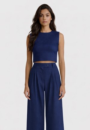 Donna con capelli lunghi, mossi e scuri, che indossa un top corto senza maniche blu navy e pantaloni larghi a vita alta blu navy, in piedi davanti a uno sfondo semplice.