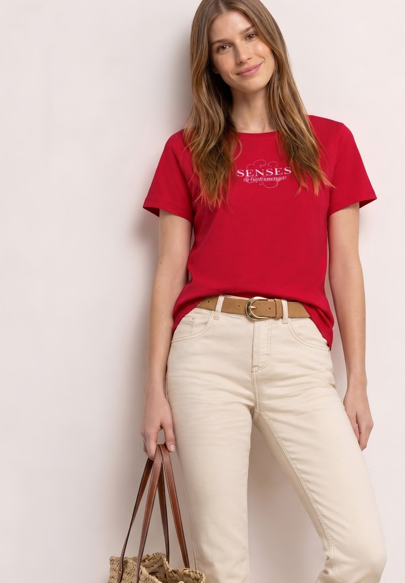 Jeune femme en t-shirt rouge imprimé et pantalon beige tenant un sac cabas tressé avec des anses en cuir, debout devant un fond neutre.