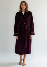 Accappatoio in morbido velluto viola intenso con cintura, maniche lunghe, piping bianco a contrasto lungo il colletto e i polsini, lunghezza fino a metà polpaccio.