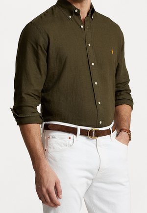 Mann trägt ein olivgrünes Hemd mit Knöpfen und hochgekrempelten Ärmeln, weiße Hose, braunen Gürtel mit goldener Schnalle und eine braune Armbanduhr, Hände in den Taschen.