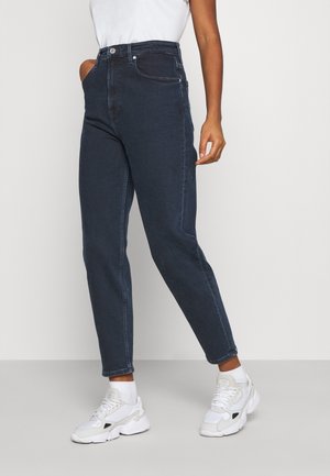 Vrouw draagt donkerblauwe high-waist jeans, witte sneakers en een wit topje, ze staat met één hand in de zak tegen een effen achtergrond.