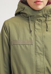 Oliwkowa kurtka z kapturem, z teksturowaną łatą na piersi, sznurkami ściągającymi oraz widocznymi detalami szycia. Gładkie wykończenie materiału.
