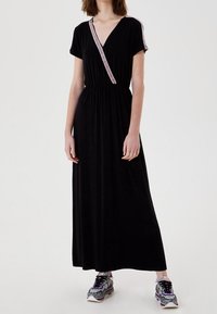 Schwarzes Maxikleid mit V-Ausschnitt, kurzen Ärmeln und einem pinkfarbenen Besatz entlang des Ausschnitts. Glatter Stoff mit elastischem Taillenbund. Kombiniert mit gemusterten Sneakers.