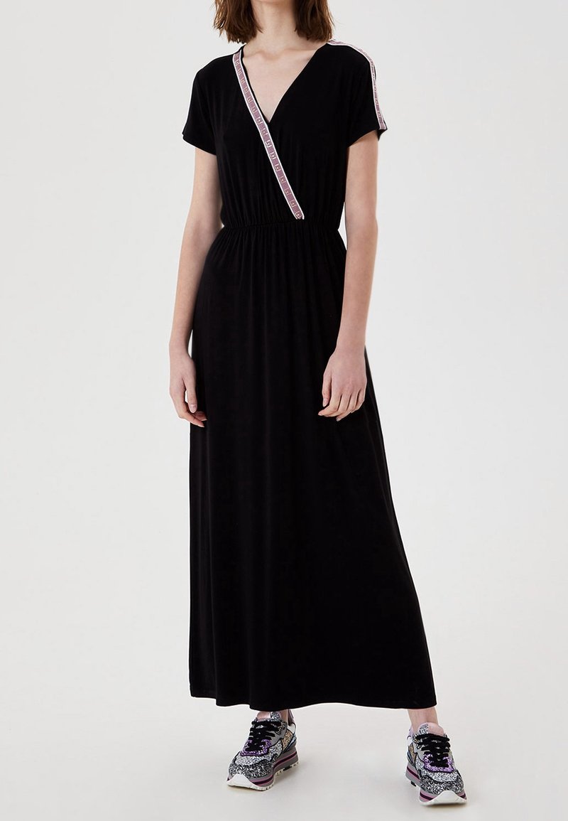 Schwarzes Maxikleid mit V-Ausschnitt, kurzen Ärmeln und einem pinkfarbenen Besatz entlang des Ausschnitts. Glatter Stoff mit elastischem Taillenbund. Kombiniert mit gemusterten Sneakers.