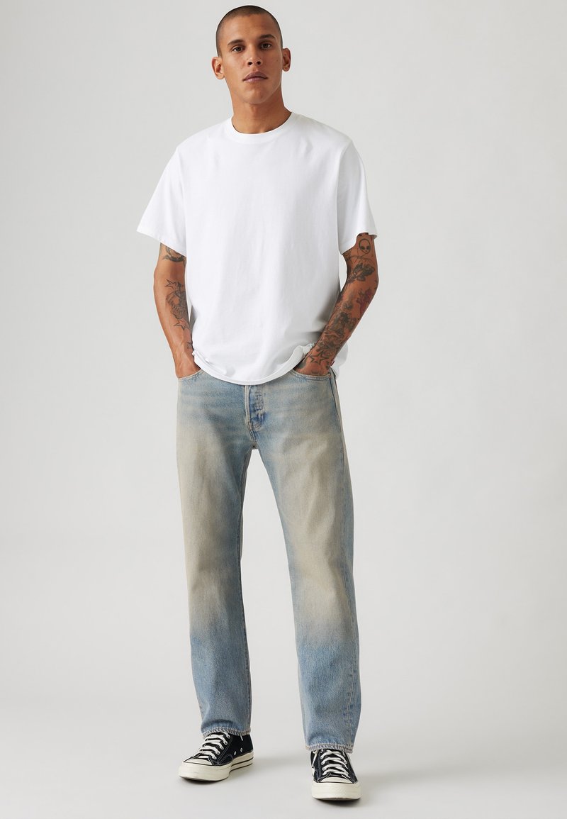Weißes Baumwoll-T-Shirt und hellblaue Jeans mit einem verblassten Waschung. Schwarze Turnschuhe mit weißen Akzenten. Tattoos auf den Armen sichtbar.