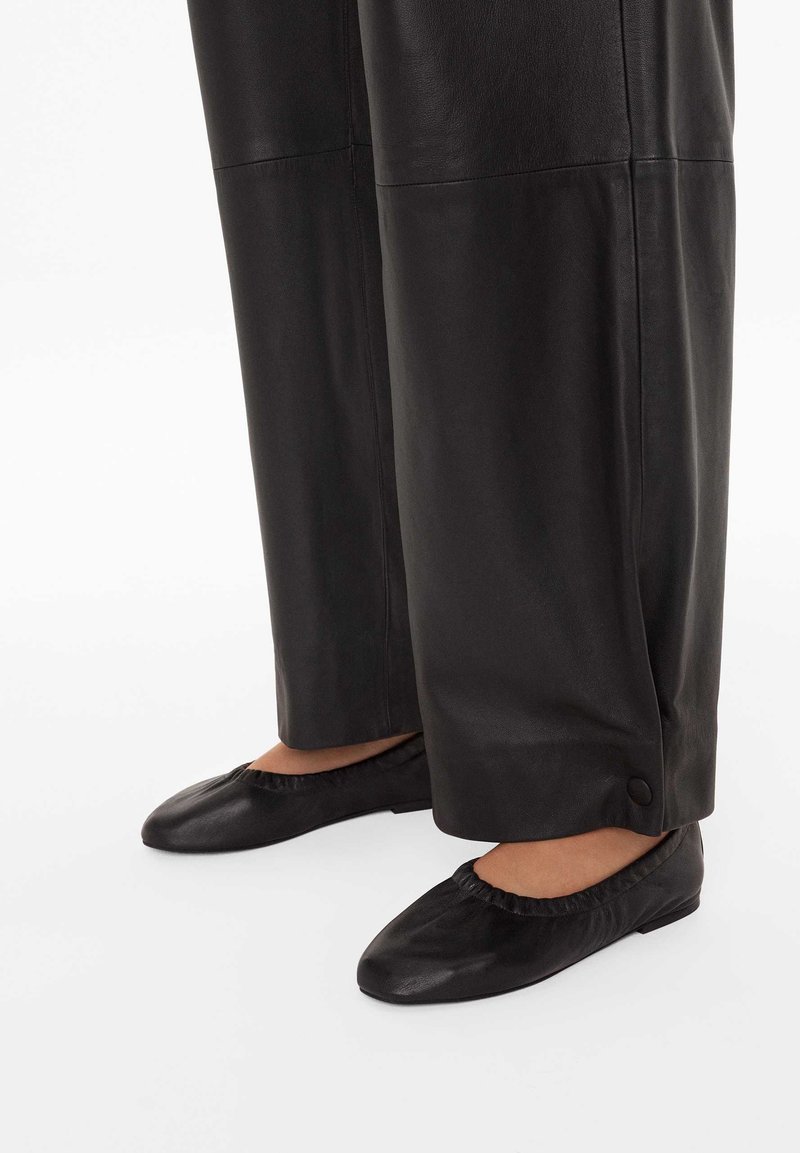 Czarne skórzane baleriny z marszczoną krawędzią i okrągłym noskiem, zestawione z szerokimi czarnymi skórzanymi spodniami z detalem guzika przy kostce.