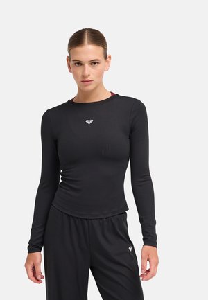 Jeune femme aux cheveux attachés porte un haut noir ajusté à manches longues et un pantalon noir ample avec de petits logos blancs sur la poitrine et la cuisse.