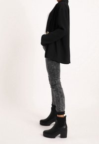 Kvinna i svart kavaj, syra-tvättade skinny jeans och svarta klackade ankelstövlar, stående mot en ren vit bakgrund.