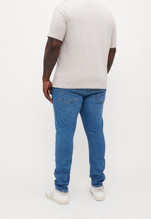 Homme portant une chemise à manches courtes beige clair, un jean bleu et des baskets blanches Veja, debout devant un fond blanc uni.