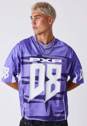 Jeune homme aux cheveux blonds courts portant un maillot de sport violet avec l'inscription "PXP 08" et des colliers en argent superposés, regardant sur le côté.