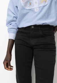 Jeans noirs à coupe slim, avec des poches avant et une fermeture à boutons, associés à une chemise rayée bleu clair avec broderie décorative.
