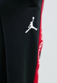 Sort stof med hvidt Jumpman-logo og en vertikal rød stribe med hvid tekst på højre side.