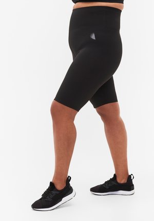 Osoba ubrana w czarne, wysokie spodenki rowerowe i czarne buty sportowe, stojąca z lekko rozstawionymi nogami na białym tle.