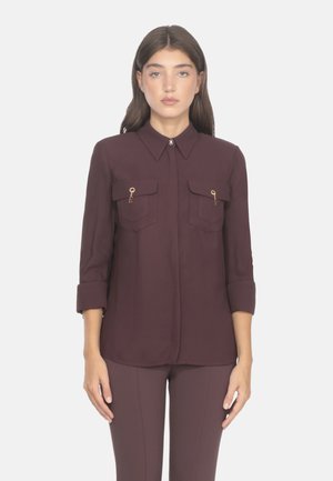 Blusa bordeaux a maniche lunghe con colletto abbottonato, due tasche sul petto e dettagli in hardware dorato. Il tessuto appare liscio e leggero.