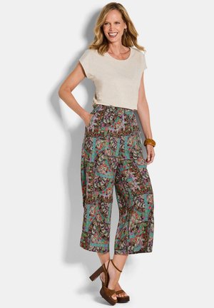 Donna sorridente con una maglietta beige a maniche corte, pantaloni larghi e cropped con motivo colorato, tacchi a zeppa marroni e un bracciale massiccio.