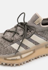 Zapatilla de punto gris con tres franjas blancas, parte superior texturizada, cordones elásticos y suela transparente con elementos de tracción texturizados.