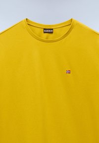 Keltainen crewneck-T-paita, jossa on pieni brodeerattu Norjan lippu vasemmalla rinnalla ja Napapijri-brändilabel kauluksessa.