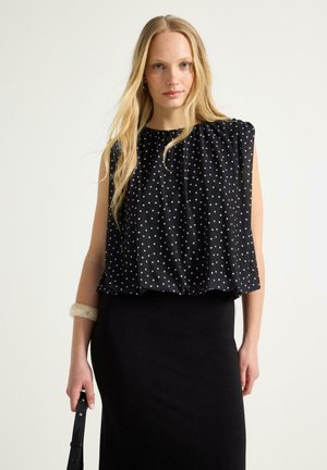 Lyshåret kvinde iført en ærmeløs sort top med hvide polkaprikker og et sort nederdel, der holder en sort rem i den ene hånd.