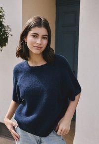 Jeune femme aux cheveux bruns mi-longs, portant des boucles d'oreilles créoles dorées, un pull court à manches courtes en laine bleu marine et un jean bleu clair, debout à l'intérieur.