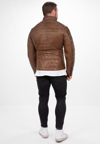 Bruine leren jas met een quiltpatroon, voorzien van een staande kraag en ritsmanchetten. Slim fit, gecombineerd met zwarte spijkerbroek en witte sneakers.