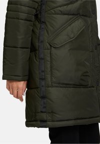 Ciemnozielona kurtka puchowa z boczną kieszenią, czarnymi wykończeniami i teksturowanym wykończeniem. Posiada zapięcie na napy oraz zamek błyskawiczny dla pewnego noszenia.