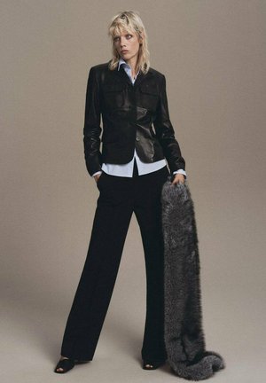 Blazer de cuero negro con dos bolsillos frontales, combinado con una camisa azul y pantalones anchos negros. Sosteniendo un chal de pelo sintético gris.