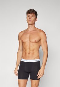 Melna bokseru apakšveļa ar baltu elastīgu jostu, uz kuras redzams "Calvin Klein" logo. Mīksta, gluda auduma, ar pielāgotu dizainu.