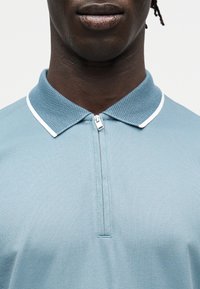 Κοντινό πλάνο ενός ατόμου που φοράει ανοιχτό μπλε πόλο μπλούζα με φερμουάρ, με υφή γιακά και λευκή επένδυση, σε απλό φόντο.