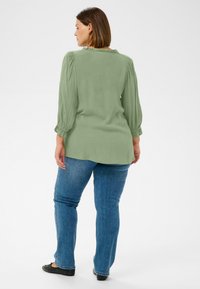 Grüne Langarmbluse mit gerafftem Schulterdetail, weicher Textur und abgerundetem Saum, kombiniert mit blauen Jeans und schwarzen Ballerinas.