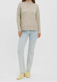 Femme portant un pull beige, un jean bleu clair et des bottines à talons jaunes, debout devant un fond uni.