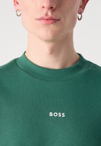 Sudadera verde con un cuello redondo acanalado y el logo blanco de "BOSS" en la parte delantera. Textura suave y silueta ajustada, que enfatiza la simplicidad.