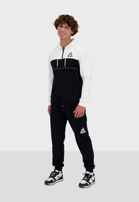 Felpa con zip bianca e nera con striscia verde, con logo sul petto; abbinata a pantaloni jogger neri, anch'essi con logo, e scarpe sportive.