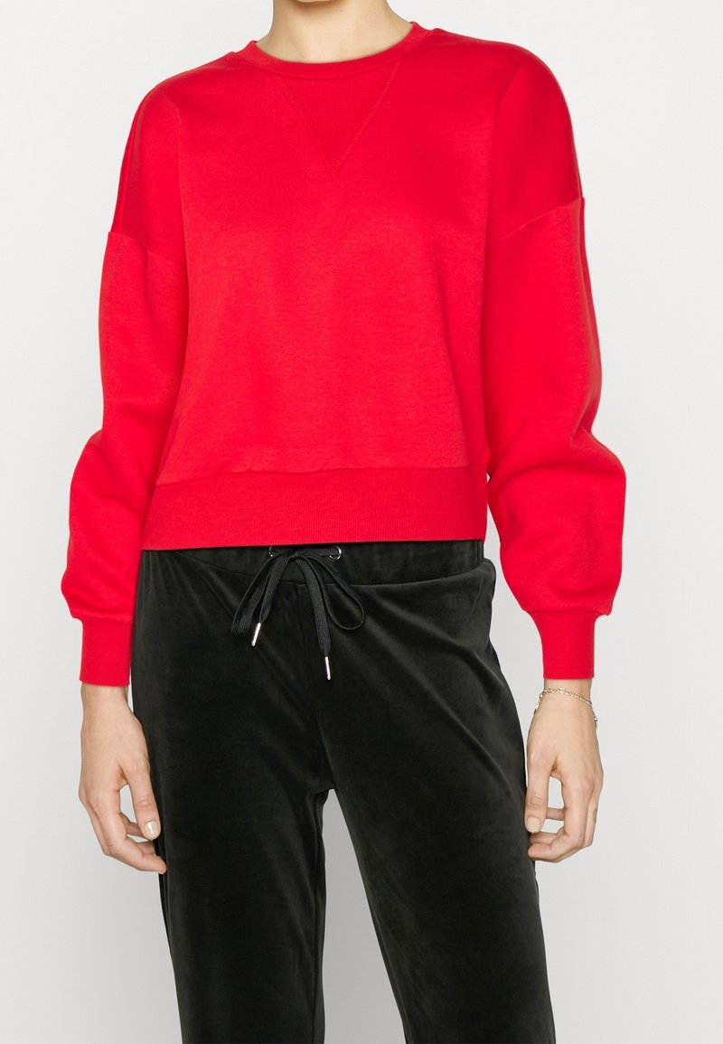 Sudadera roja recortada con cuello y puños acanalados, con hombros caídos. Combinada con pantalones de terciopelo negros y cordón negro.