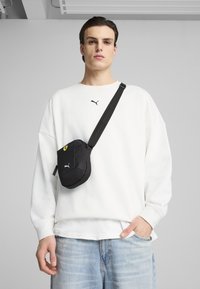 Sort crossbody-taske med Puma-logo, båret over en hvid oversized sweatshirt. Denimjeans med en lys vask og diskret udtoning.