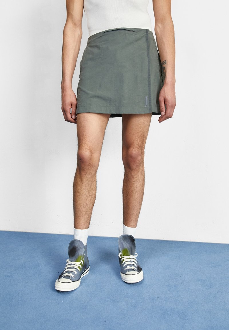 Personne portant une mini-jupe grise, des chaussettes blanches et des baskets montantes bicolores, debout sur un sol bleu contre un mur blanc.