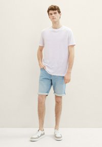 Camiseta blanca de manga corta, pantalones cortos de mezclilla azul claro con los bordes enrollados, zapatillas blancas y grises, de pie sobre un fondo neutro claro.