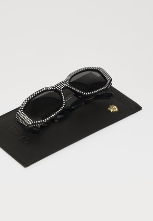Black sunglasses with a hexagonal shape, embellished with rhinestones along the frame, placed on a black leather case with a gold emblem.