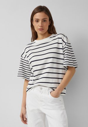 Kobieta ubrana w białą koszulkę z krótkim rękawem z czarnymi poziomymi paskami oraz białe spodnie, pozująca z jedną ręką w kieszeni na szarym tle.